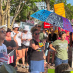 A crowd of demonstrators. One holds an umbrella, on which is written "No Kings" and "Only Queens".