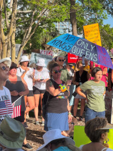 A crowd of demonstrators. One holds an umbrella, on which is written "No Kings" and "Only Queens".