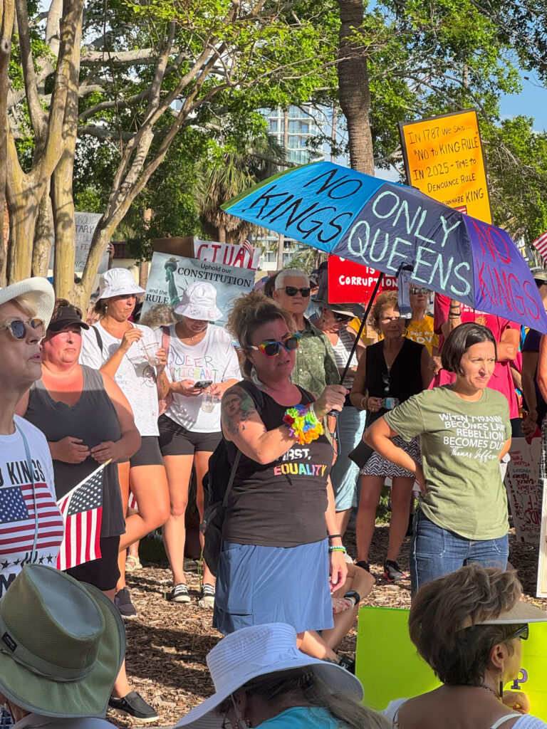 A crowd of demonstrators. One holds an umbrella, on which is written "No Kings" and "Only Queens".