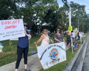 Demonstrators along the sidewalk hold signs, including one that reads "Boycott Avelo; no deportations without die process!"