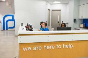 Receptionists at a desk with a sign that reads "we are here to help." The Planned Parenthood logo is visible on a wall in the background.