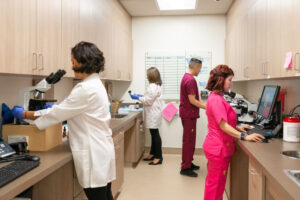 Staff at a medical facility. One professional looks at a computer. Another operates a microscope.