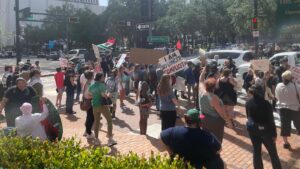 A crowd of protesters. One had a pro-Palestine sign that reads "From the river to the sea." Another's sign reads "I am no longer complicit." Another's reads "Abolish ICE." Across the street is a cluster of pro-Trump counter protesters.