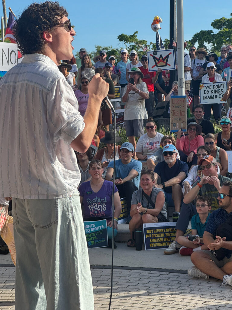 Shot from behind speaker Zander Moricz as he addresses a large crowd holding signs with messages such as "We don't do kings in America" and wearing shirts with messages such as "Protect trans kids."