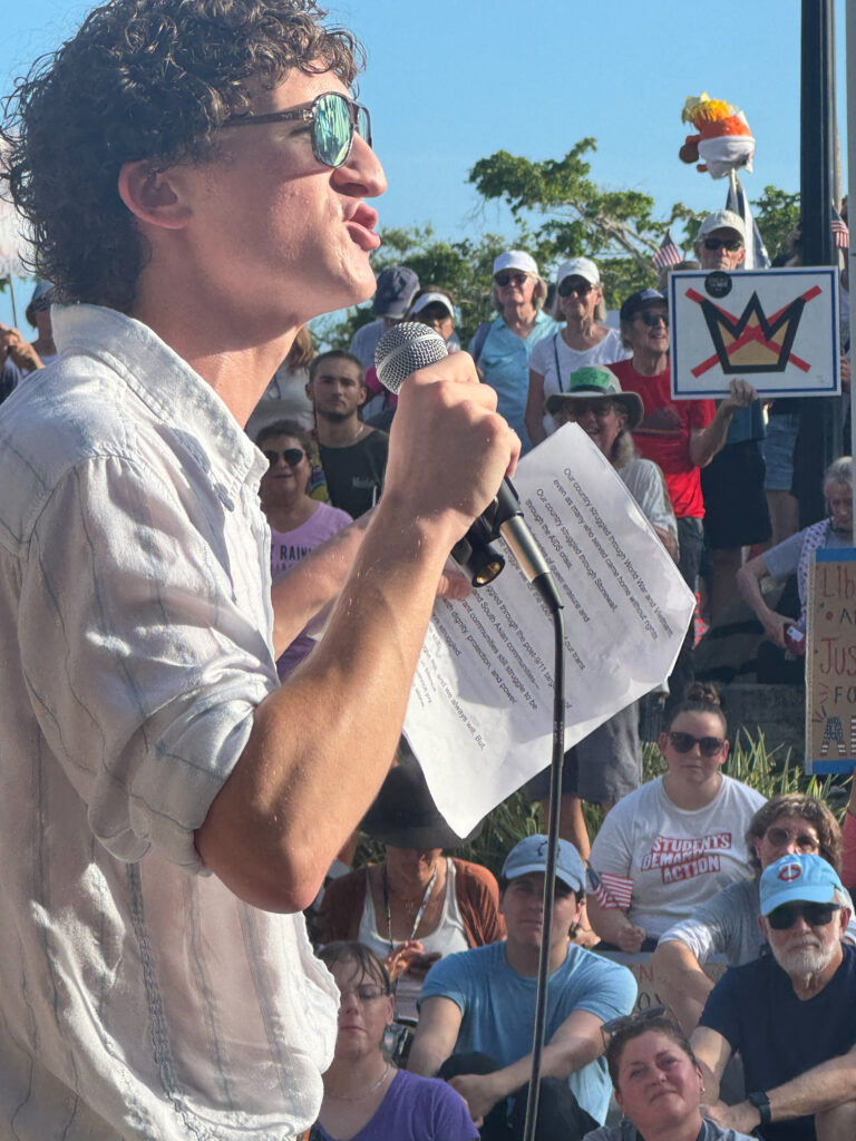 Shot from behind speaker Zander Moricz as he addresses a large crowd. One protester holds a sign featuring the No Kings logo, a crown with an "X" through it.