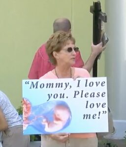 A person holding a sign featuring an illustration of a fetus with the text "Mommy, I love you. Please love me!"