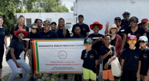 Photo of a group of kids and adults smiling as they present a banner that reads "Manasota ASALH Freedom School: Black history classes for kindergarten to 12th grade students" with the Manasota ASALH logo and a border with the Pan-African colors.