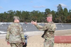 Two people in camo uniforms stand on a beach, one gesticulating while speaking.