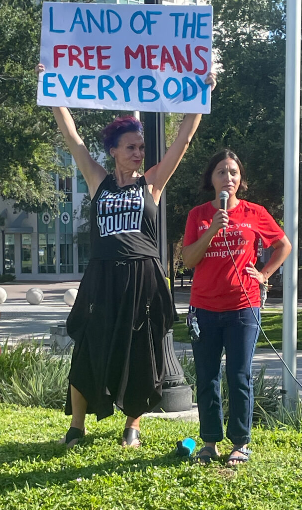 India X holding a sign that reads "Land of the free means everybody" and wearing a tank top that reads "Protect trans youth," standing next to Julie Forrestier holding a microphone and wearing a shirt that says "I want for you whatever you want for immigrants".