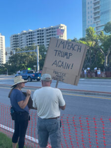 Two demonstrators photographed from behind. One holds a sign that reads, "Impeach Trump again! Practice makes perfect".