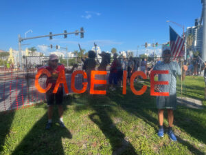 Two protesters hold up a sign that appears to be made out of a segment of fence with text that reads "Cage ICE".