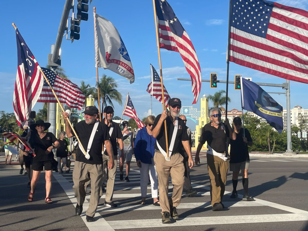 A group of protesters wearing matching shirts and holding flags for the U.S. and its armed forces cross the street.