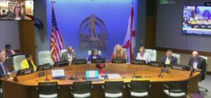 Commissioners meeting at the Sarasota City Commission Hall arouynd a conference table with the City of Sarasota logo behind them. Liz Alpert is visible on a video call in the top left.