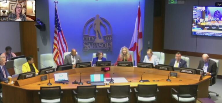 Commissioners meeting at the Sarasota City Commission Hall arouynd a conference table with the City of Sarasota logo behind them. Liz Alpert is visible on a video call in the top left.