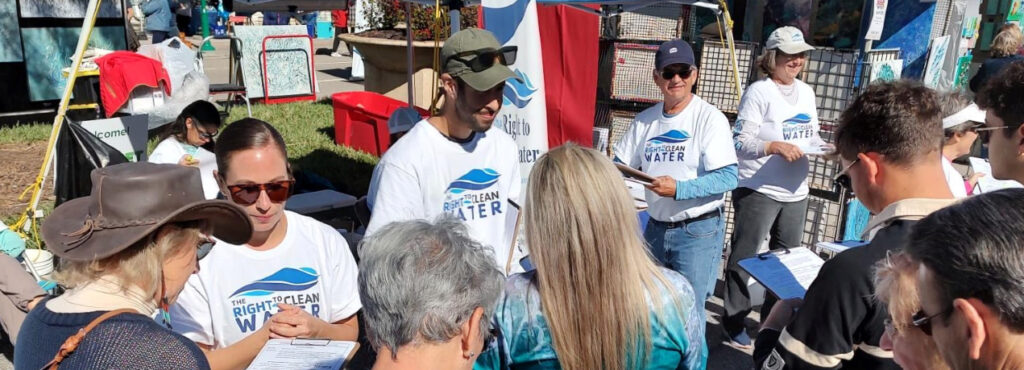 A crowd approaches a group of volunteers wearing T-shirts that say "The Right to Clean Water" at an outdoor convention.