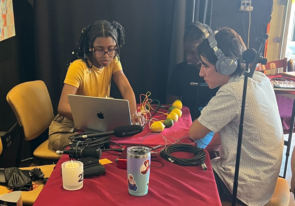 Three young people regarding a laptop. On the table nearby, wires appear to be plugged into several lemons and limes.