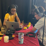 Three young people regarding a laptop. On the table nearby, wires appear to be plugged into several lemons and limes.