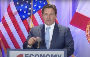 Ron Desantis gesticulating while speaking behind a podium in front of U.S. flag and Florida state flag.