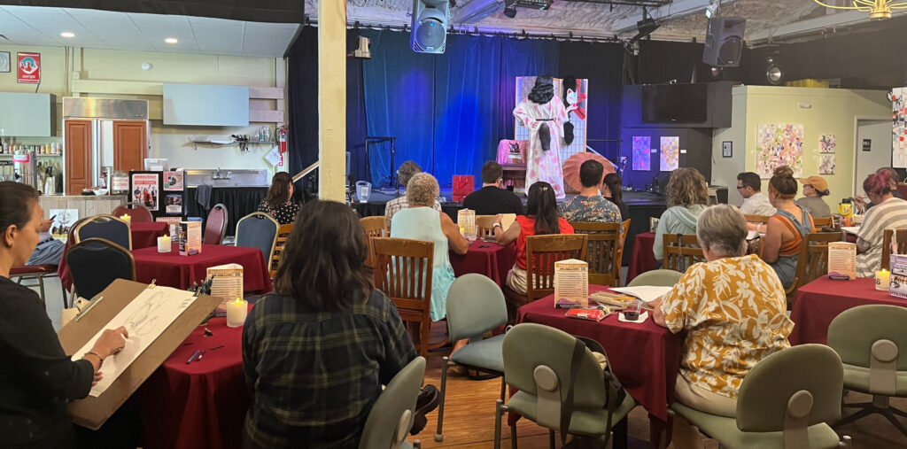 A crowd of artists with sketchbooks and drawing boards draw a model onstage at Fogartyville.