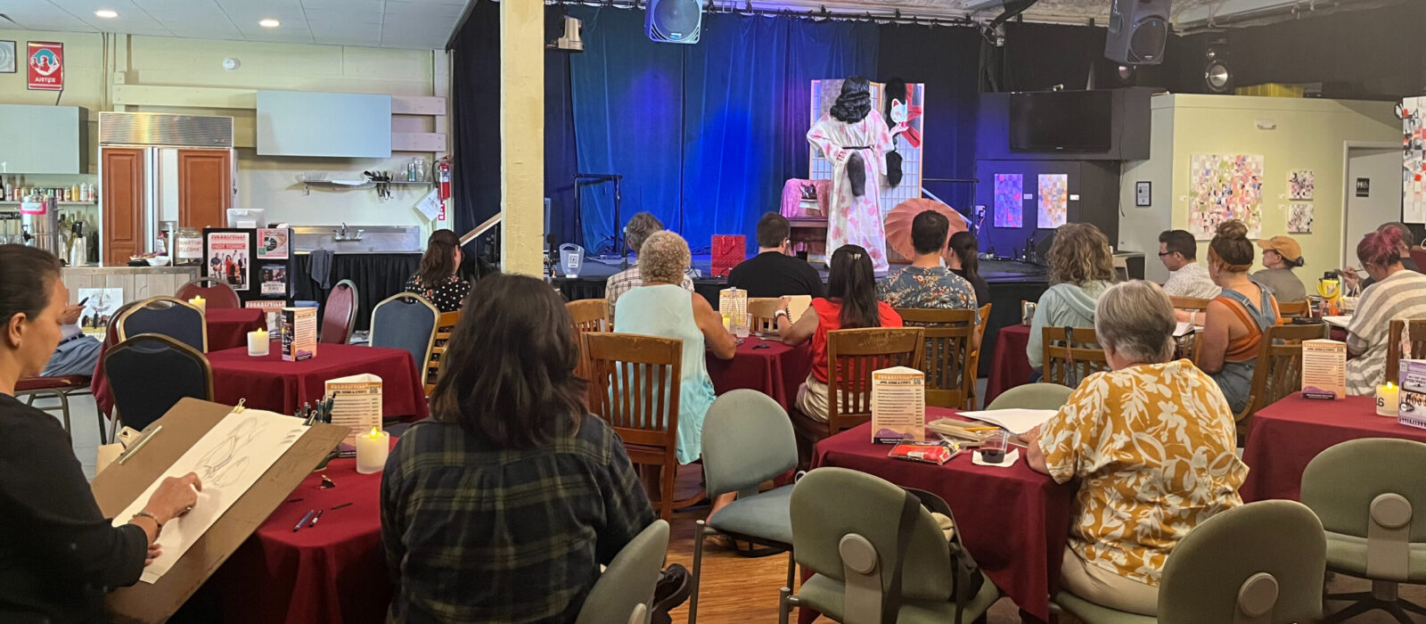 A crowd of artists with sketchbooks and drawing boards draw a model onstage at Fogartyville.