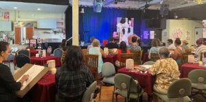 A crowd of artists with sketchbooks and drawing boards draw a model onstage at Fogartyville.