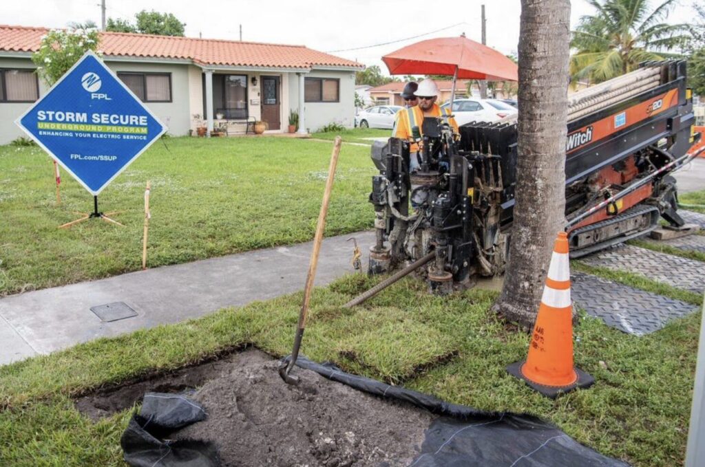 Workers in hard hats and hi-vis vests standing by a shallow hole freshly dug in the ground. An FPL-branded sign nearby reads, "Storm Secure Underground Program: Enhancing your electric service / FPL.com/SSUP".