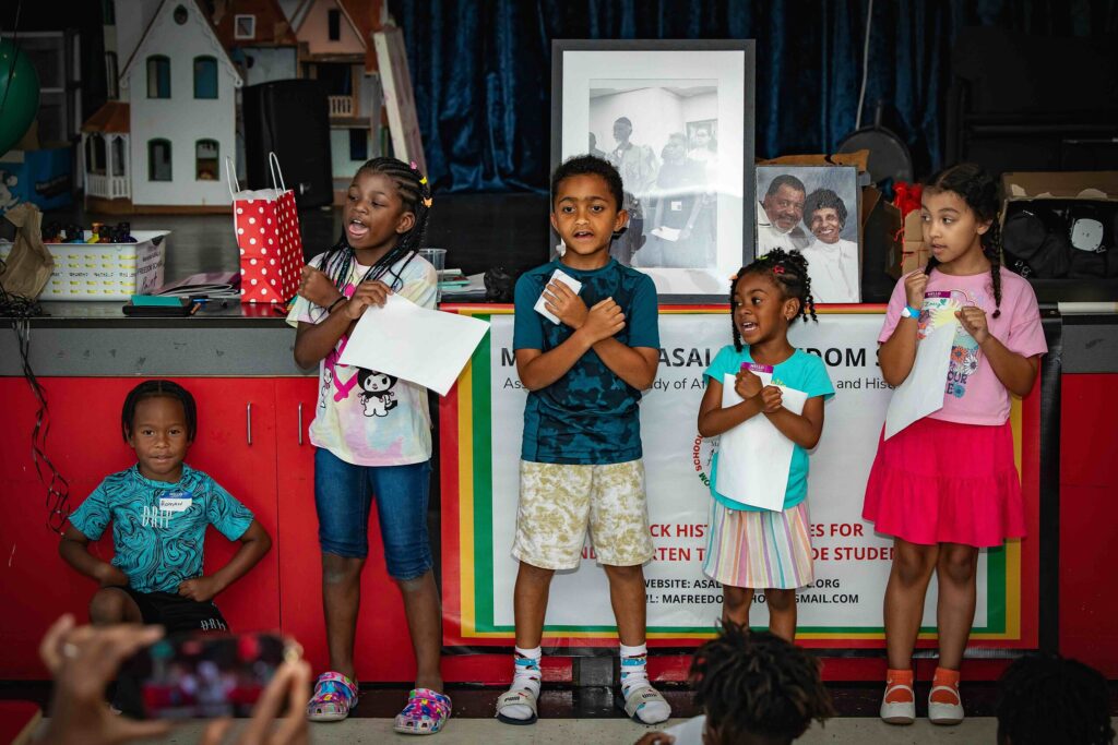Five children perform in front of a crowd/ The three in the middle have their arms crossed over their chests.