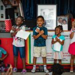Five children perform in front of a crowd/ The three in the middle have their arms crossed over their chests.