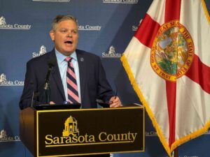 Jonathan Lewis speaking at a podium in front of a Florida state flag.
