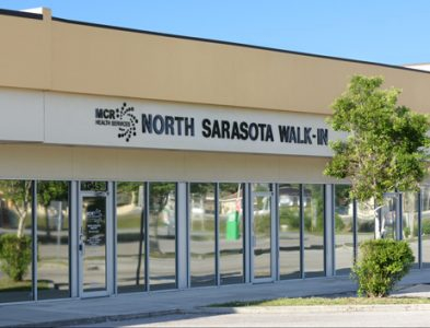 The front of a building. The sign features a logo for MCR Health Services next to text that reads "North Sarasota Walk-In."