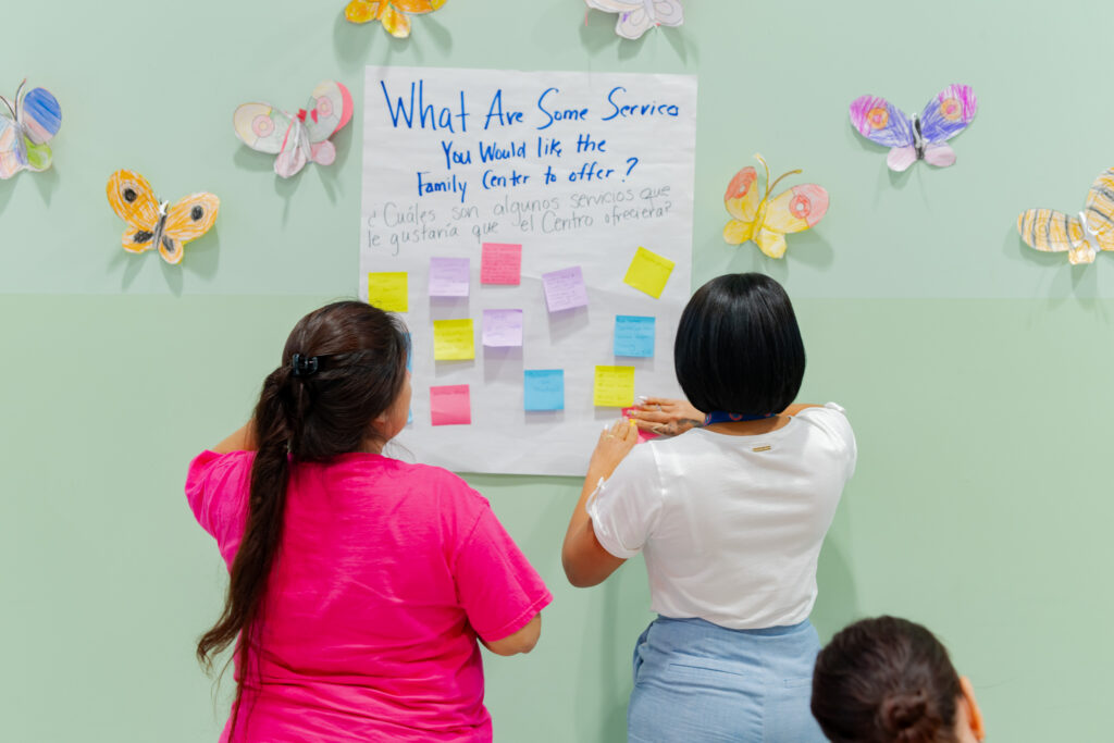 Two people affix sticky notes to a wishlist on a wall, upon which is written "What are some services you would like the Family Center to offer? / ¿Cuáles son algunos servicios que la gustaría que el Centro ofreciera?". Paper butterflies surround the wishlist.