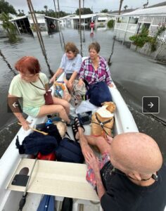 A group of people and a dog pack a small boat floating atop a flooded neighborhood street.