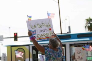 A demonstrator, viewed from behind, holds a sign that says "We the People means everybody" with a trans and gay pride flag drawn on it.