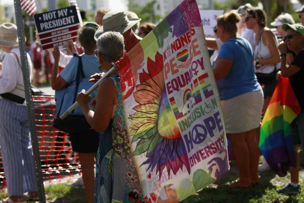 Demonstrators hold signs outdoors. One in the foreground holds a pride flag with a floral design which reads "Kindness, Peace, Equality, Love, Inclusion, Hope, Diversity."