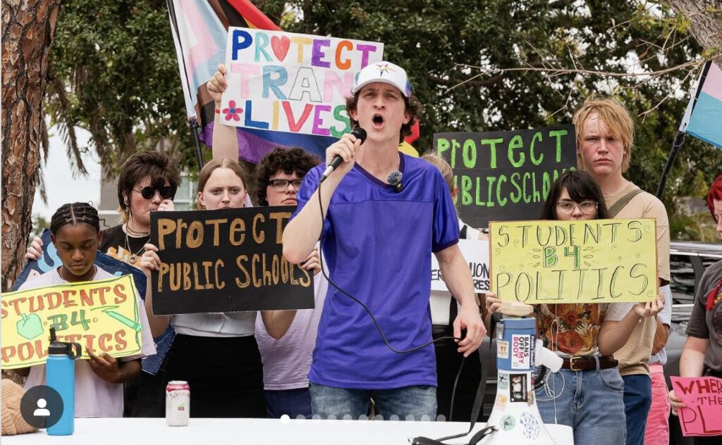 Zander Moricz speaks passionately into a microphone in front of demonstrators holding handwritten signs that read "protect trans lives," "protect public schools," and "students B4 politics."