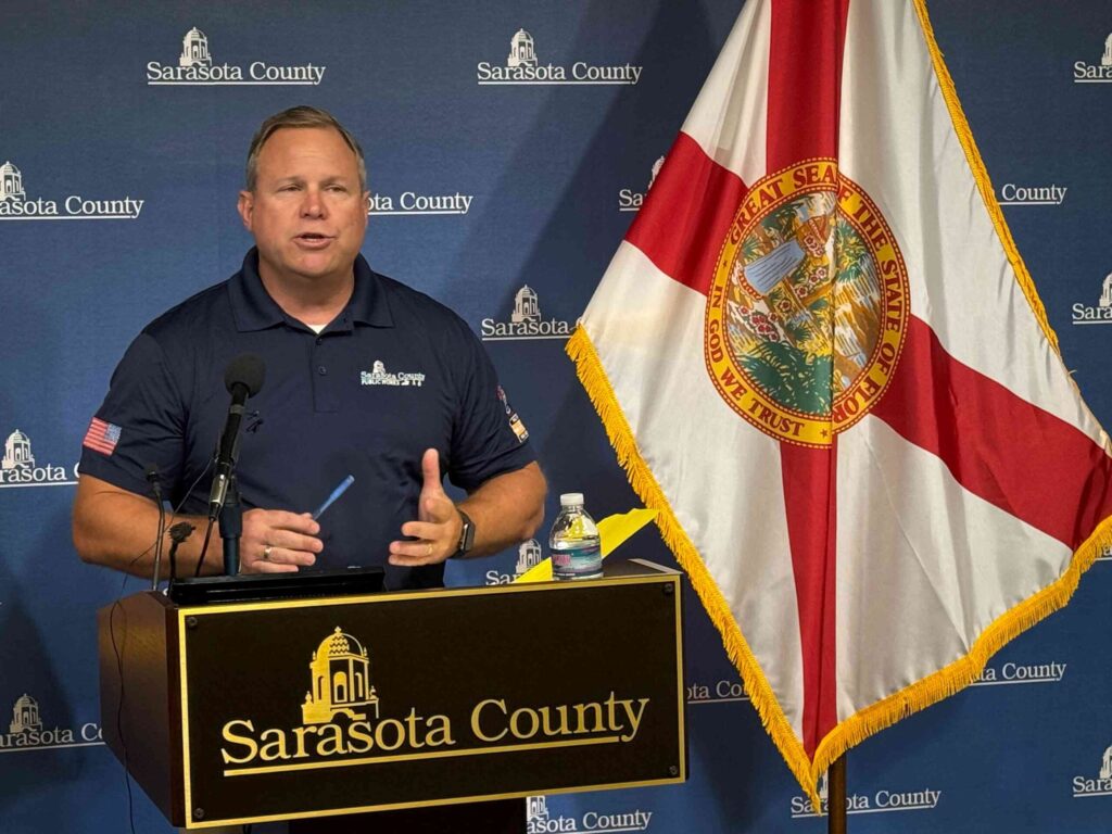 Spencer Anderson speaking at a podium in front of a Florida state flag.
