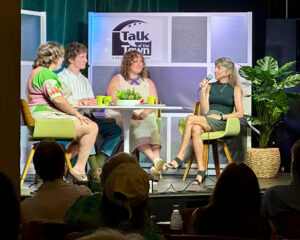 Panelists sit around a table in front of a crowd. Behind them is the Talk of the Town: A WSLR News Roundtable logo.