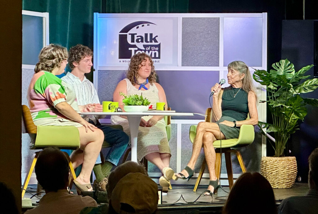 Panelists sit around a table in front of a crowd. Behind them is the Talk of the Town: A WSLR News Roundtable logo.