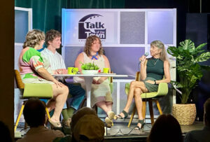 Panelists sit around a table in front of a crowd. Behind them is the Talk of the Town: A WSLR News Roundtable logo.