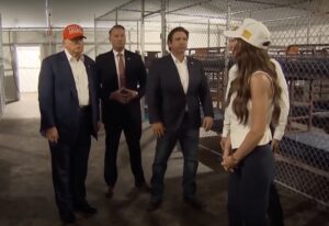 President Donald Trump, Gov. Ron DeSantis, and other stand inside a tent near bunk beds enclosed with chain-link fences.