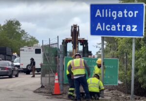Workers in hi-vis clothing put up a blue roadside sign that reads "Alligator Alcatraz."