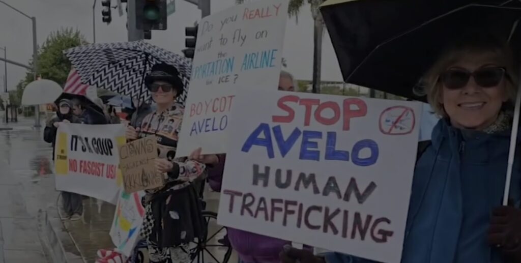 Demonstrators outside in the rain hold signs with messages including "Stop Avelo human trafficking", "Do you really want to fly on the deportation airline for ICE? Boycott Avelo" and "It's a coup; no Trump; No fascist USA".