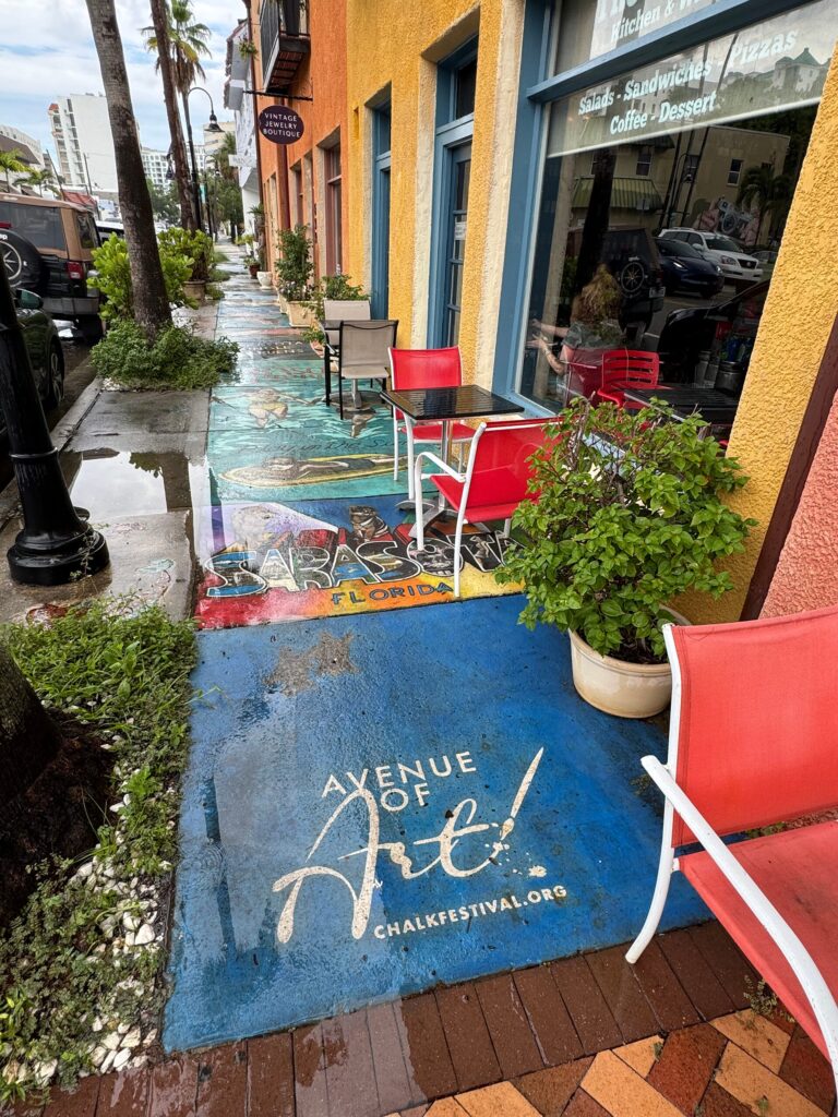 Photo of a sidewalk adjacent to a line of buildings downtown. Each section of the sidewalk is painted with a different design. The closest one features text that reads "Avenue of Art! chalkfestival.org".