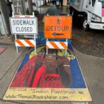 Sidewalk painting of a person with peacock feathers in their hair wearing an ornate red garment and holding a paintbrush, partially obscured by road work signs atop the subject's face.