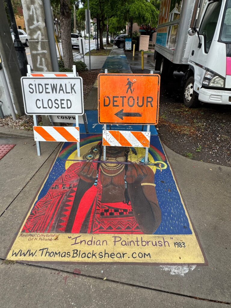 Sidewalk painting of a person with peacock feathers in their hair wearing an ornate red garment and holding a paintbrush, partially obscured by road work signs atop the subject's face.