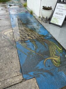 Sidewalk painting of a fish jumping out of the sea. The fish breaks the border created by the sidewalk grooves, creating a 3D effect.