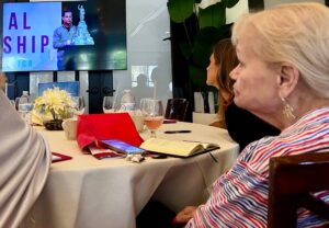 Attendees at a table watching a presenter on stage and on screen. Partially visible text behind the presenter appears to read: -AL -SHIP