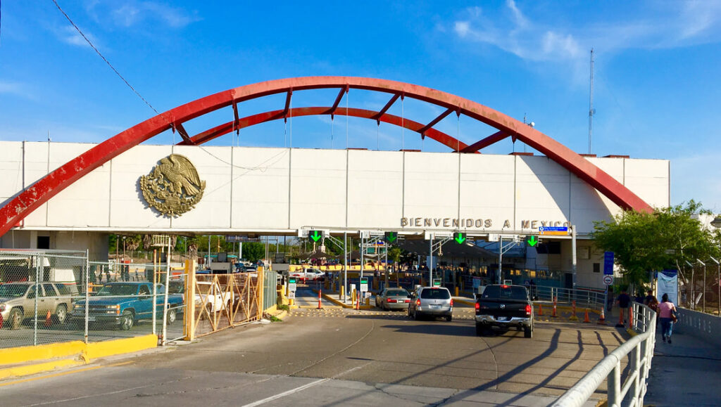 Cars drive through a border crossing area with a decorative red bridgelike arch and a sign that reads "Bienvenidos a Mexico."
