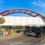 Cars drive through a border crossing area with a decorative red bridgelike arch and a sign that reads "Bienvenidos a Mexico."