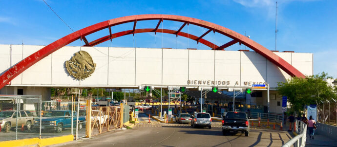 Cars drive through a border crossing area with a decorative red bridgelike arch and a sign that reads "Bienvenidos a Mexico."